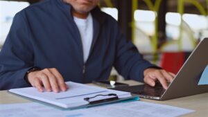 Person working at a laptop while reviewing paperwork and documents spread across a table.